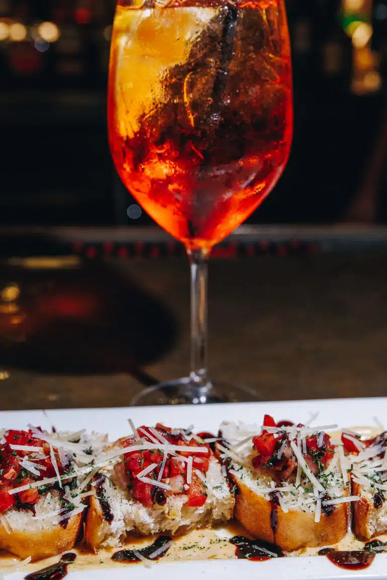 A wine glass filled with a bright orange colored Aperol spritz behind a plate of toasted bruschetta.