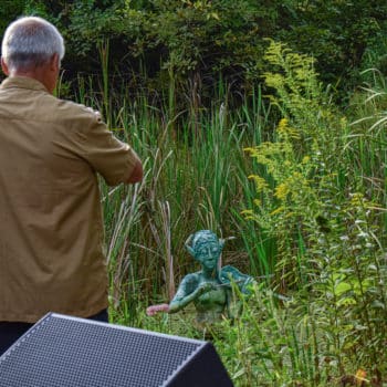 Man taking a picture of a sculpture in the pond. The sculpture is a green, somewhat fishlike, person.