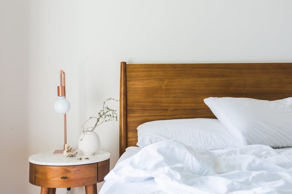 unmade bed with whit comfy sheets and comforter. bedside table with flower and dainty lamp