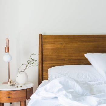 unmade bed with whit comfy sheets and comforter. bedside table with flower and dainty lamp
