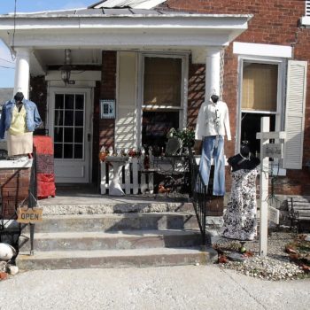 exterior shot of older brick home with items for sale displayed on the porch, steps, and sidewalk. Ms. Mary, the Mannequin, in the foreground