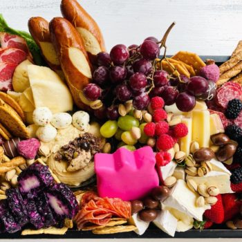 A charcuterie board with grapes, cheese, raspberries, pretzels, salami, crackers, chocolate covered almonds, pistachios, and more.