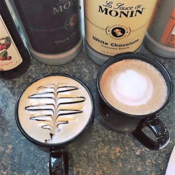 two cups of coffee, with designs cut in with cream, setting in front of a few flavored syrup bottles